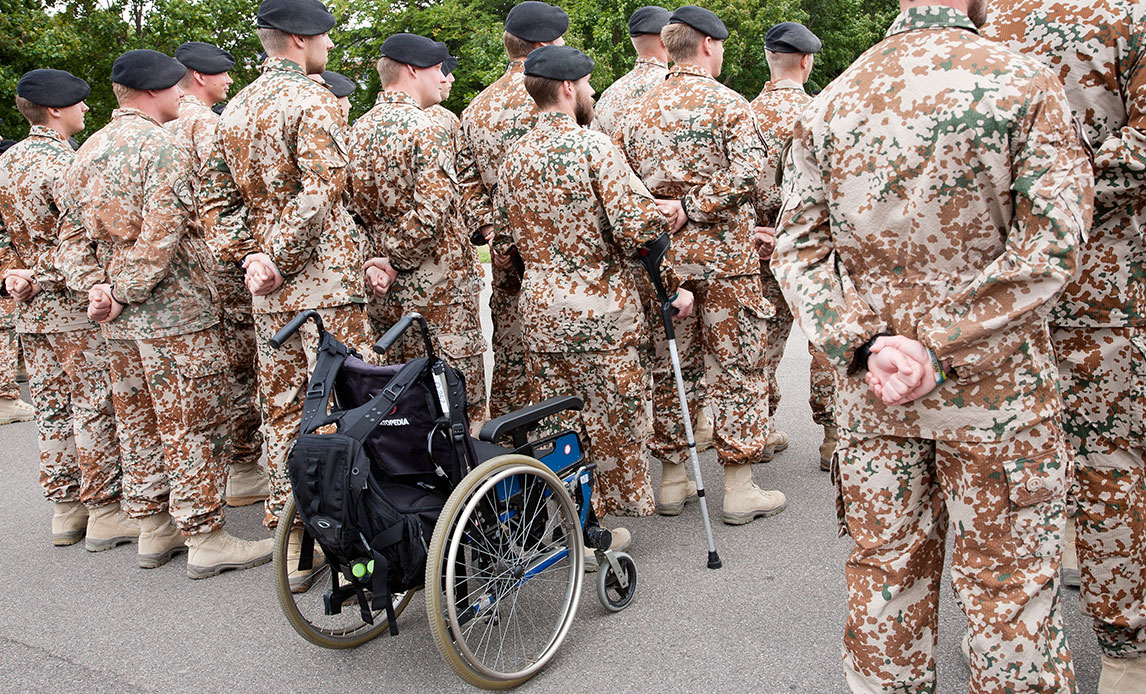 En gruppe af uniformerede soldater står med ryggen til, en enkelt står med støtte fra krykker og med sin kørestol bag sig.
