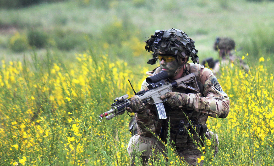 Mandlig soldat med sløring i ansigtet, hjelm og uniform sidder på hug og sigter med sit gevær i en gul rapsmark.